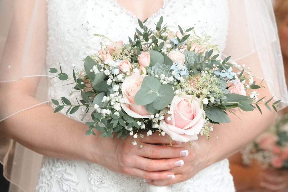 A bride holding a delicate bouquet arrangement featuring soft pink roses, pale blue hydrangeas, sprigs of white baby's breath, and eucalyptus leaves, wrapped with subtle white ribbon. The bouquet is c