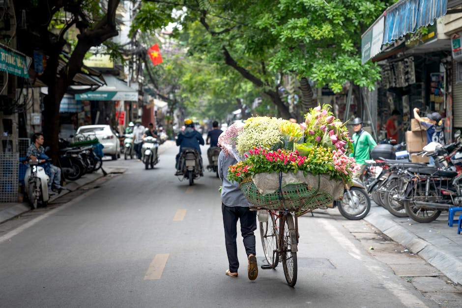A street scene showing a person pushing a bicycle loaded with a large, colorful floral bouquet featuring pink, white, green, and yellow flowers, including roses, carnations, and hydrangeas, wrapped pa