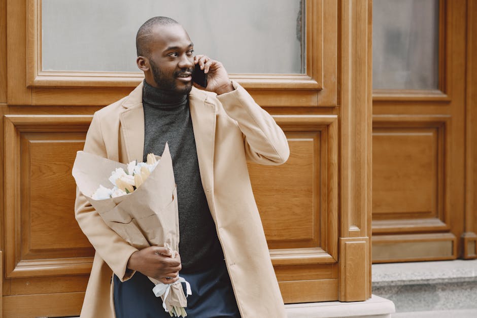 A man with dark skin, a neatly trimmed beard, and a cheerful expression is standing outdoors against a wooden-paneled building, holding a bouquet of fresh cream and white flowers wrapped in brown craf