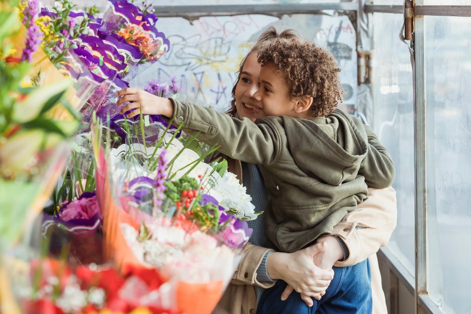 A young boy with curly hair and a smile, wearing a green hooded jacket, is being held by an adult with light skin, in front of a display of vibrant floral bouquets. The boy is reaching out to touch th
