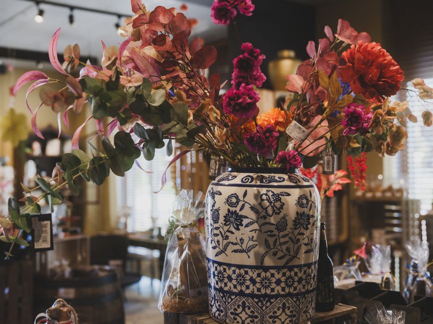 A large floral arrangement in a decorative ceramic vase featuring blue floral patterns on a white background, placed on a wooden surface inside a flower shop. The arrangement includes vibrant pink and