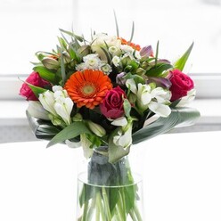 This stunning bouquet, thoughtfully arranged by expert hands in Feltham, is a masterful display of balance, colour, and artisan skill. The floral symphony begins with four prominent gerbera daisies-two in dazzling orange that evoke the vibrance of Feltham's bustling High Street, and two in silken peach, their creamy petals glowing with understated elegance around bold, deep brown centers. Interwoven are rich magenta roses, which add depth and an aura of sophistication reminiscent of Feltham's traditional rose gardens. White alstroemeria and clusters of petite spray roses provide a crisp, clean lift to the arrangement, each petal luminous under the gentle wash of morning light. Subtle touches of pale pink and fine white filler blooms nestle among the leaves, producing a sense of richness and abundance that mirrors the varied textures and colours of Feltham Park in spring. Anchoring the bouquet are broad, glossy aspidistra leaves, carefully shaped around the base for support, while smaller pointed leaves and gentle green sprigs weave throughout, building contrast and fullness. The arrangement sits in a sleek, clear glass vase, through which vibrant stems are seen beneath the water's surface-a nod to freshness and attention to quality. The effect is a visual and sensory delight, suited to anniversaries, housewarmings, or moments of gratitude. Each petal and leaf is a testament to the joy and optimism found in Feltham, lovingly curated for lasting impact.